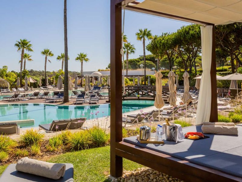 A resort swimming pool area with lounge chairs, palm trees, and a covered cabana with drinks on a daybed.