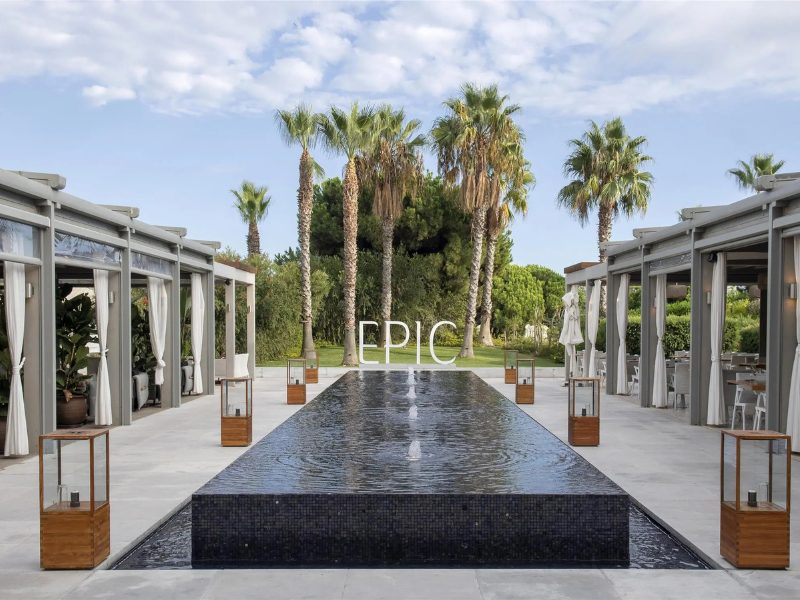 A long rectangular water feature leads to large white letters spelling "EPIC" in front of palm trees and lush greenery. White-curtained cabanas with dining tables line the walkway on either side.