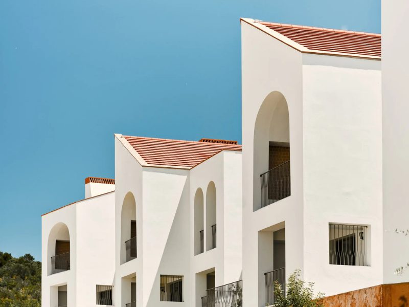 White buildings with arched doorways and windows, topped with terracotta roofs, against a bright blue sky.
