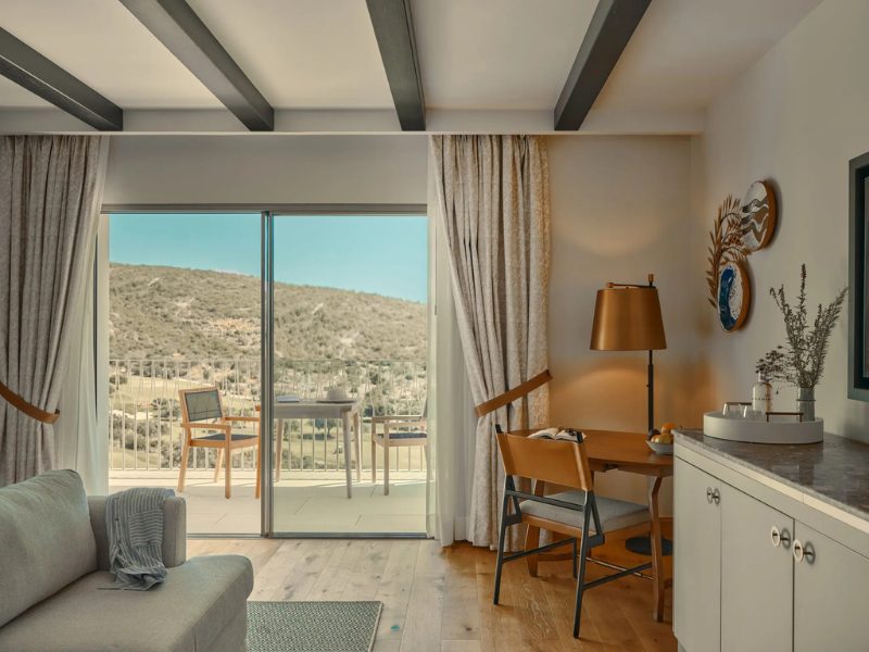 A living room with a sofa, a desk with a lamp, and a balcony overlooking a hilly landscape.
